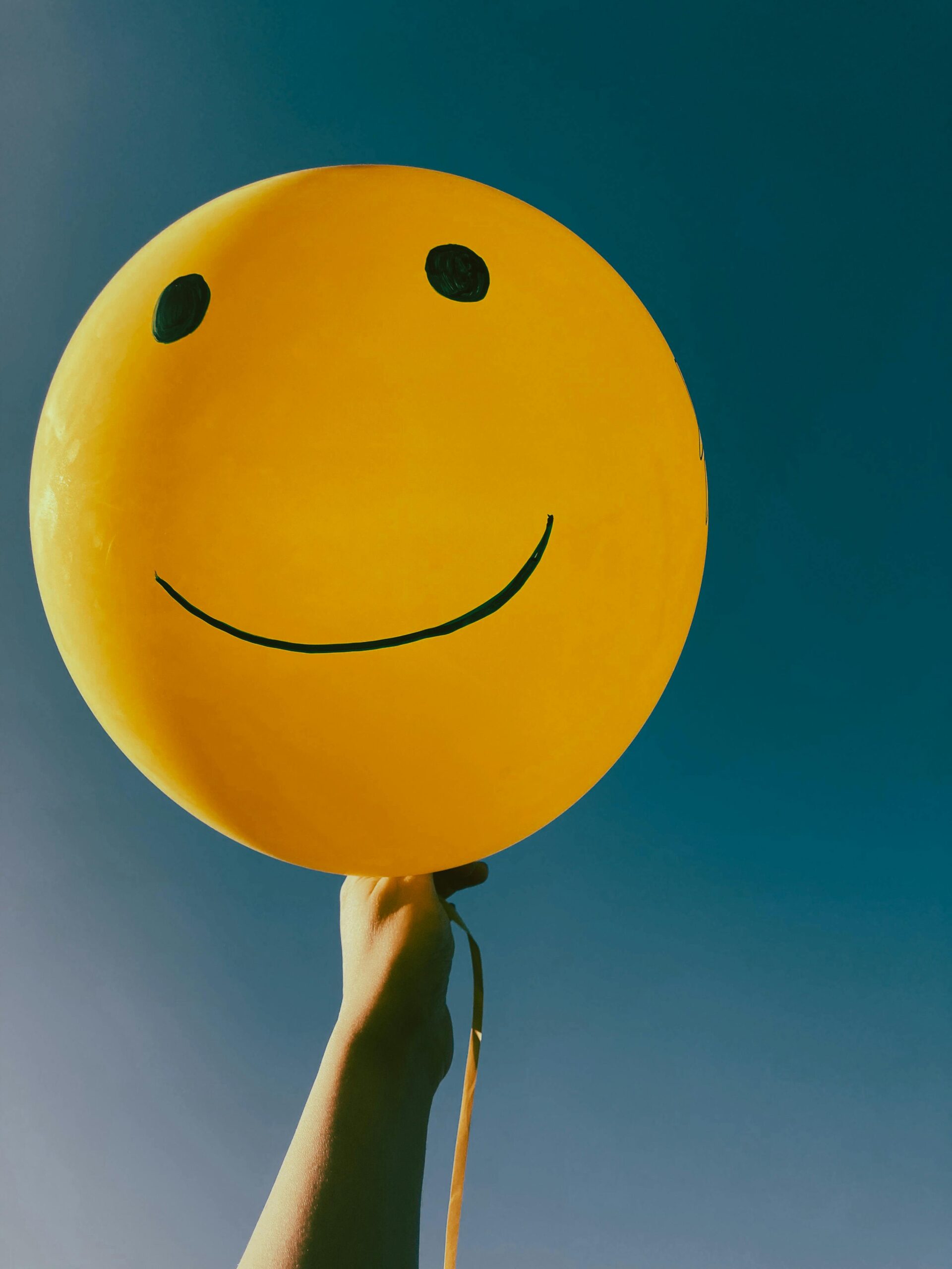 Bright yellow smiley balloon held aloft against a clear blue sky, evoking happiness and positivity.
